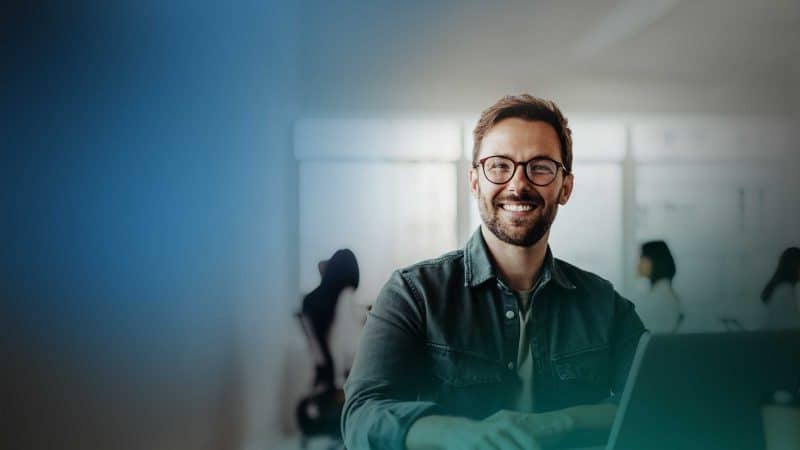 Ein junger, bärtiger Mann mit Brille sitzt lachend vor seinem Laptop.