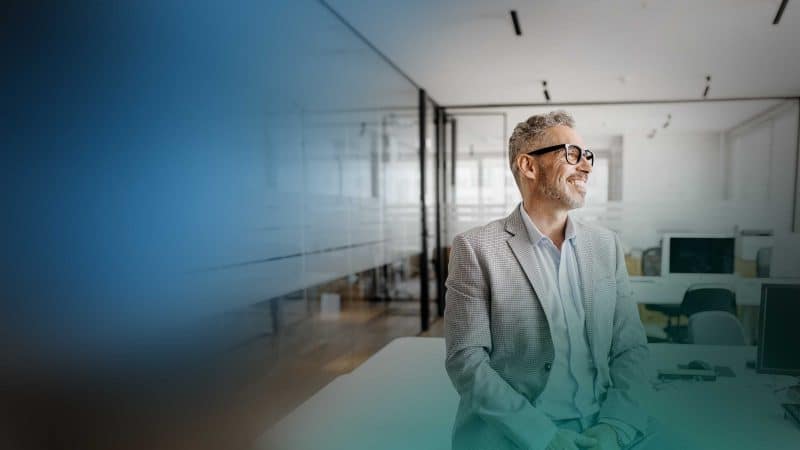 Ein Mann in hellem Anzug steht in einem Großraumbüro an einer Scheibe und blickt hinaus.
