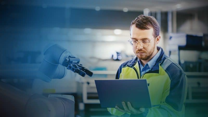 Ein Mann mit gelber Arbeitsweste steh in einem technischen Labor und schaut auf seinen Laptop.