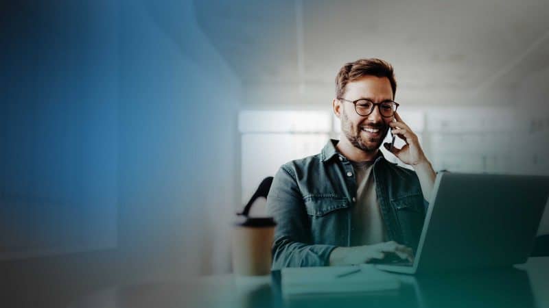 Mann sitzt am Laptop und telefoniert mit einem Smartphone.