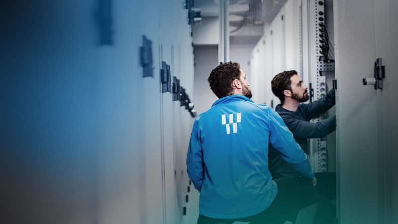 Das Bild zeigt zwei Männer, die vor einem Cage in einem Rechenzentrum hocken.