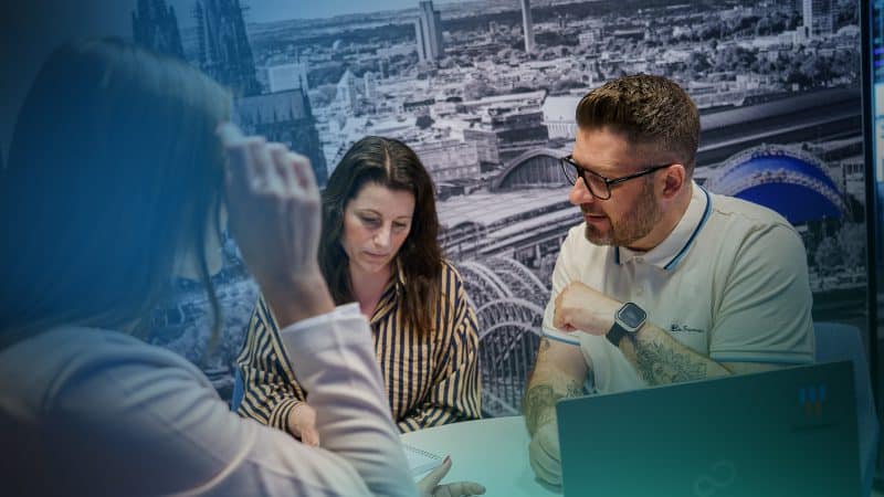 Drei Personen sitzen in einem Meeting, im hintergrund ist das Panorama der Stadt Köln mit Dom zu sehen